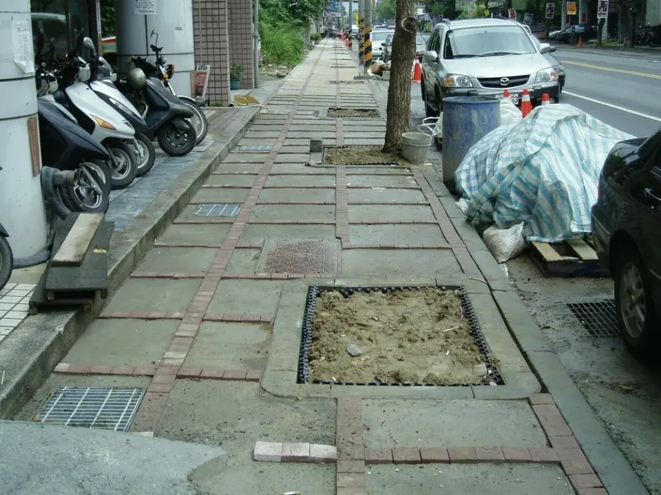 磚砌人行道上的方形樹穴,土面外圍有黑色框格,旁有機車與路邊車輛